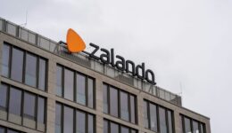 A modern building with large windows features a sign for Zalando, including its logo—an orange, rounded triangle—mounted on the rooftop against an overcast sky. The sign is in bold, black letters.