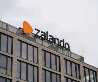 A modern building with large windows features a sign for Zalando, including its logo—an orange, rounded triangle—mounted on the rooftop against an overcast sky. The sign is in bold, black letters.
